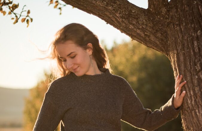 Standing outside in the sun and leaning her left hand against a tree young woman with long brown hair faces us, smiling slightly. She wears a brown sweater, her eyes closed and face directed away from us, towards the ground next to her.
