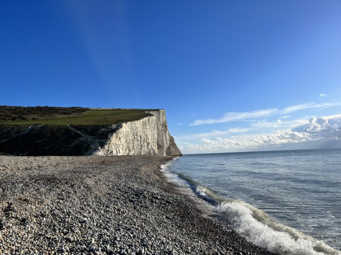 Under a bright blue sky, a pebble beach is lapped at by the sea. Both stretch into the distance towards tall white cliffs covered by green vegetation.
