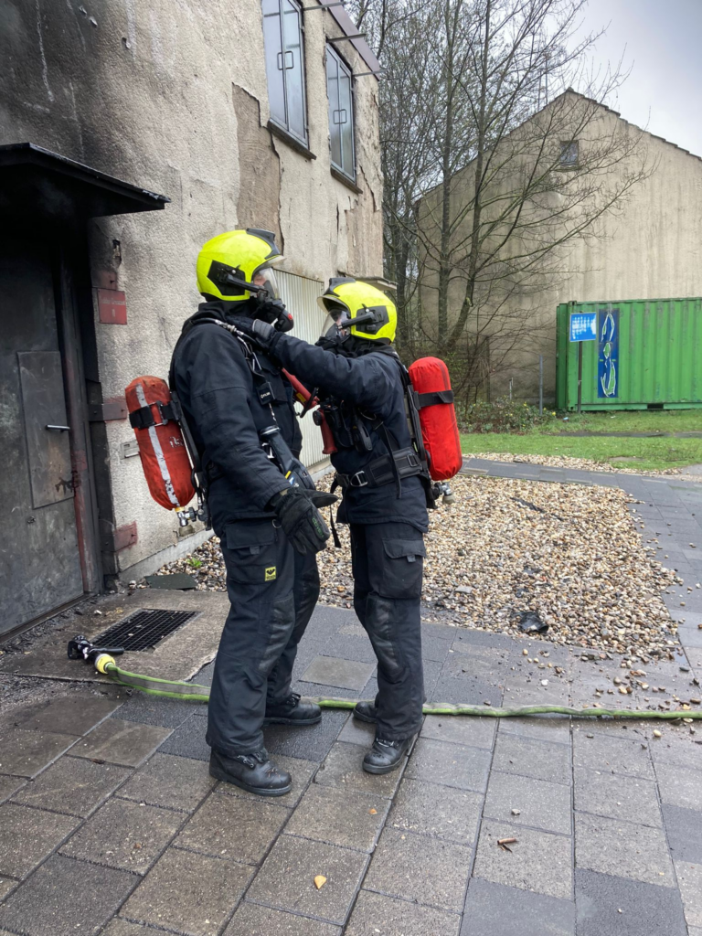 Two firefighters in black fire suits and neon green helmets are standing outside a brick building's metal door, facing each other. They are wearing breathing gear and red air bottles on their backs. One of the firefighters is holding the helmet flap of the other, checking for possible exposed skin. On the tiled ground is a green firehose. In the far background another building can be seen, a green shipping container standing in front of it.
